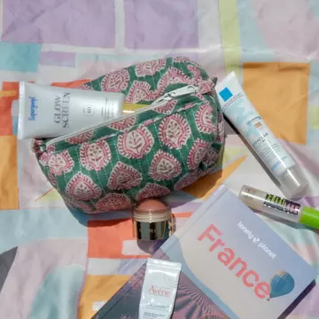 A patterned pouch with a zipper on a patterned scarf with a Lonely Planet guidebook for France and makeup products.