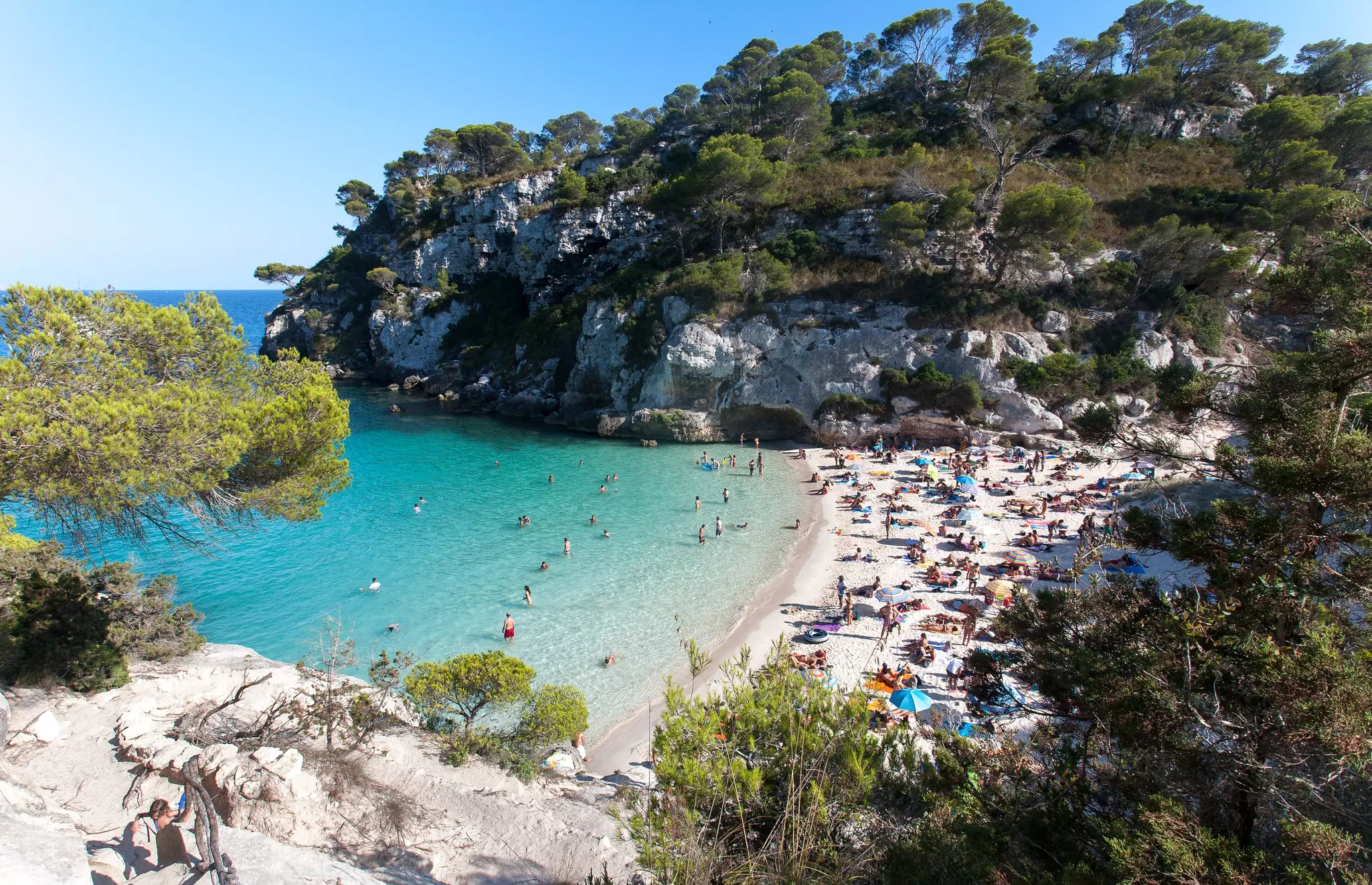 A wide view of a cove with a small beach framed by a limestone headland, and with bright blue water