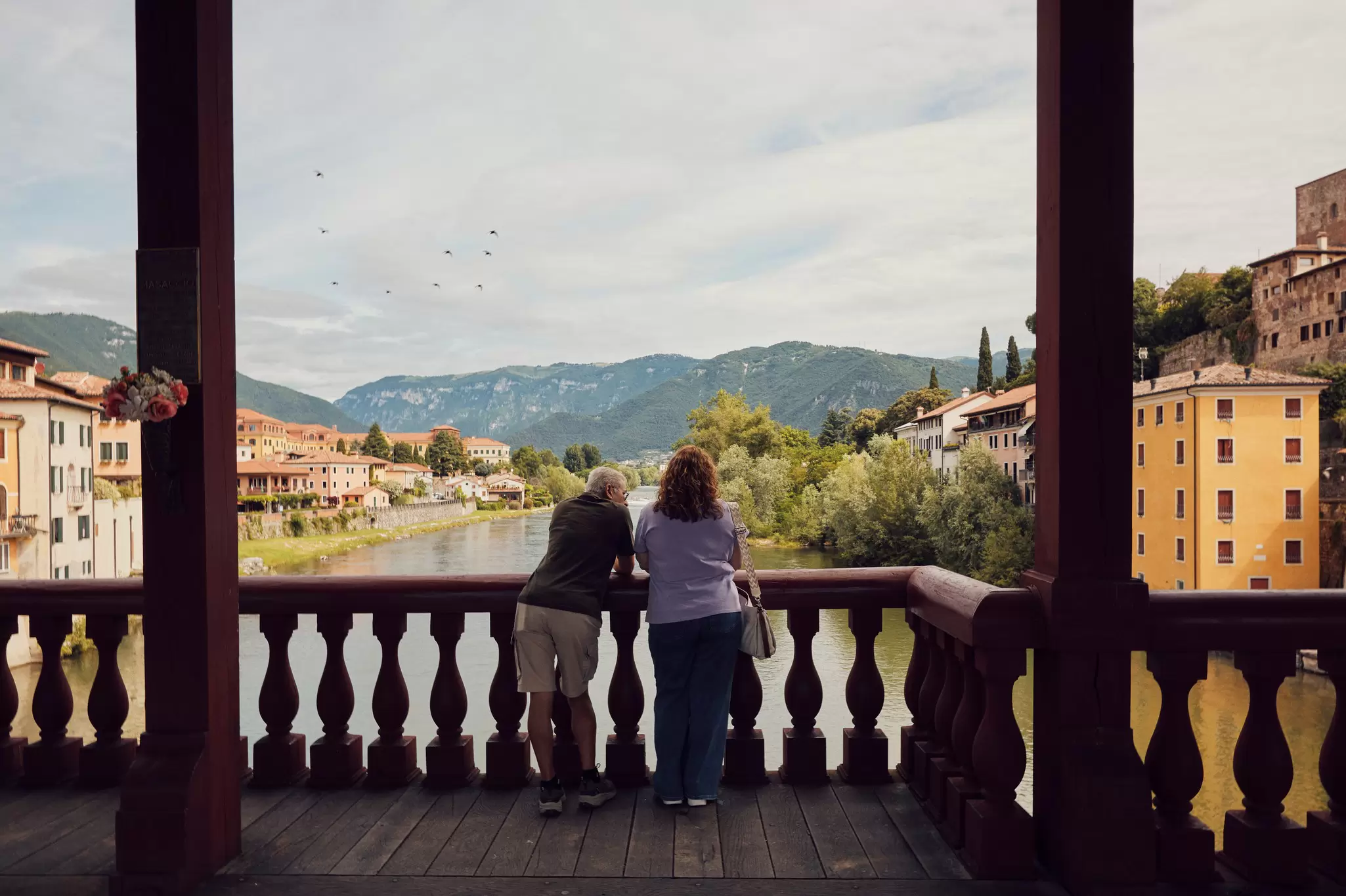 A couple stands on a bridge overlooking a river, historic city, and mountains