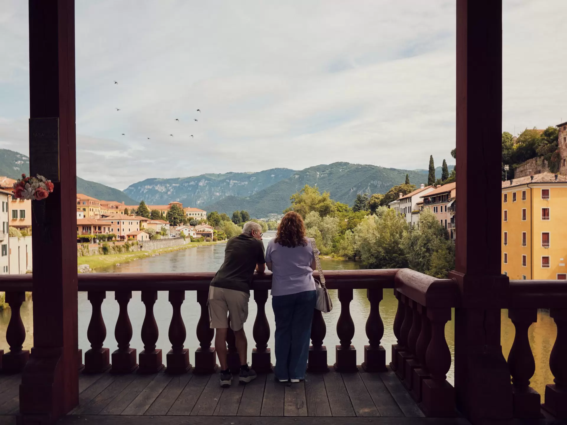 A couple stands on a bridge overlooking a river, historic city, and mountains
