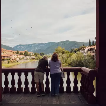 A couple stands on a bridge overlooking a river, historic city, and mountains