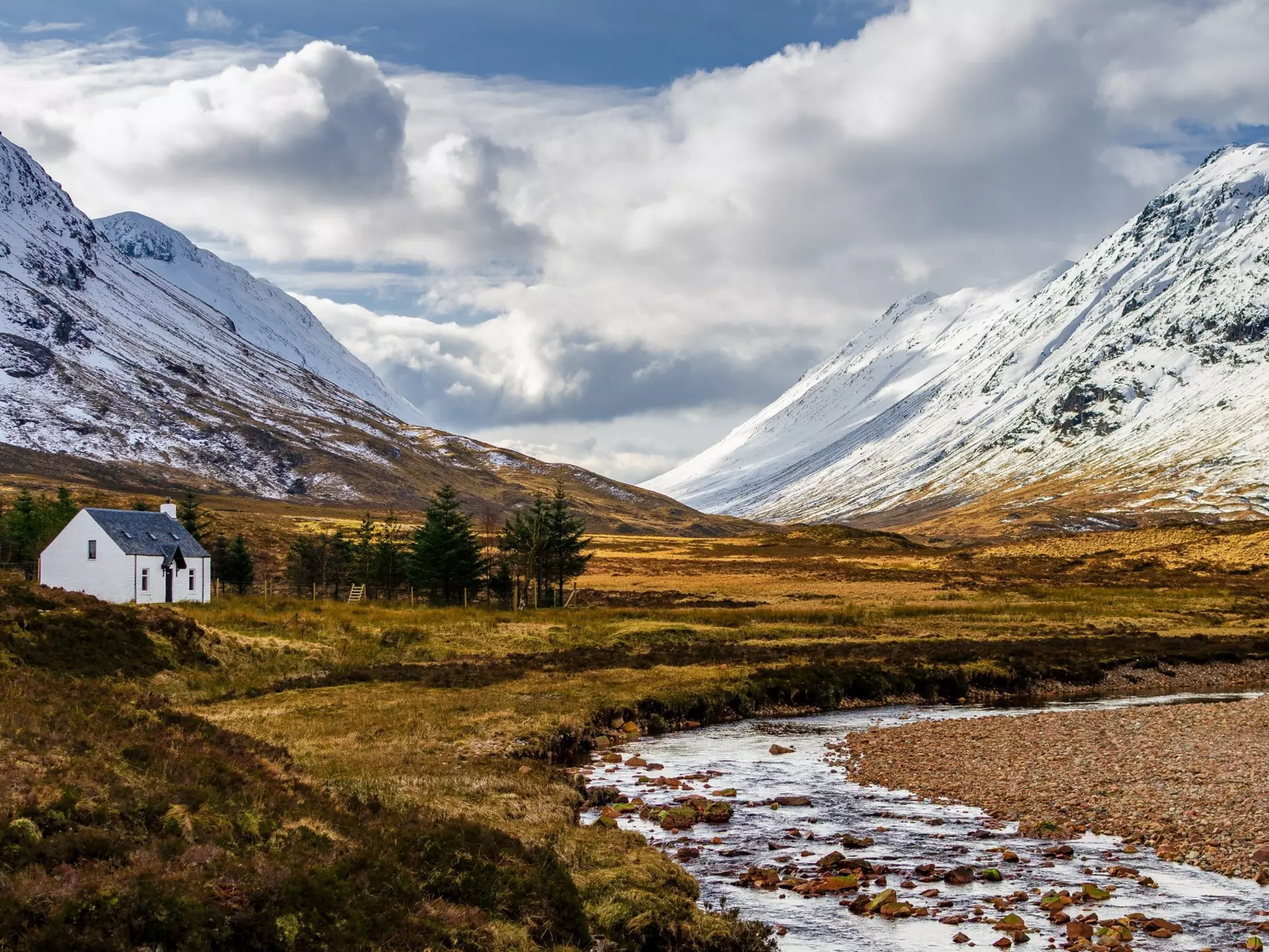 A stone cottage stands near a small stream running through a valley surrounded by snow-capped hills in Glencoe, Scotland.