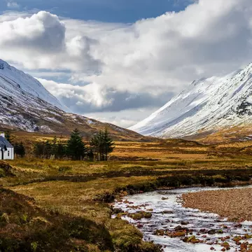 A stone cottage stands near a small stream running through a valley surrounded by snow-capped hills in Glencoe, Scotland.