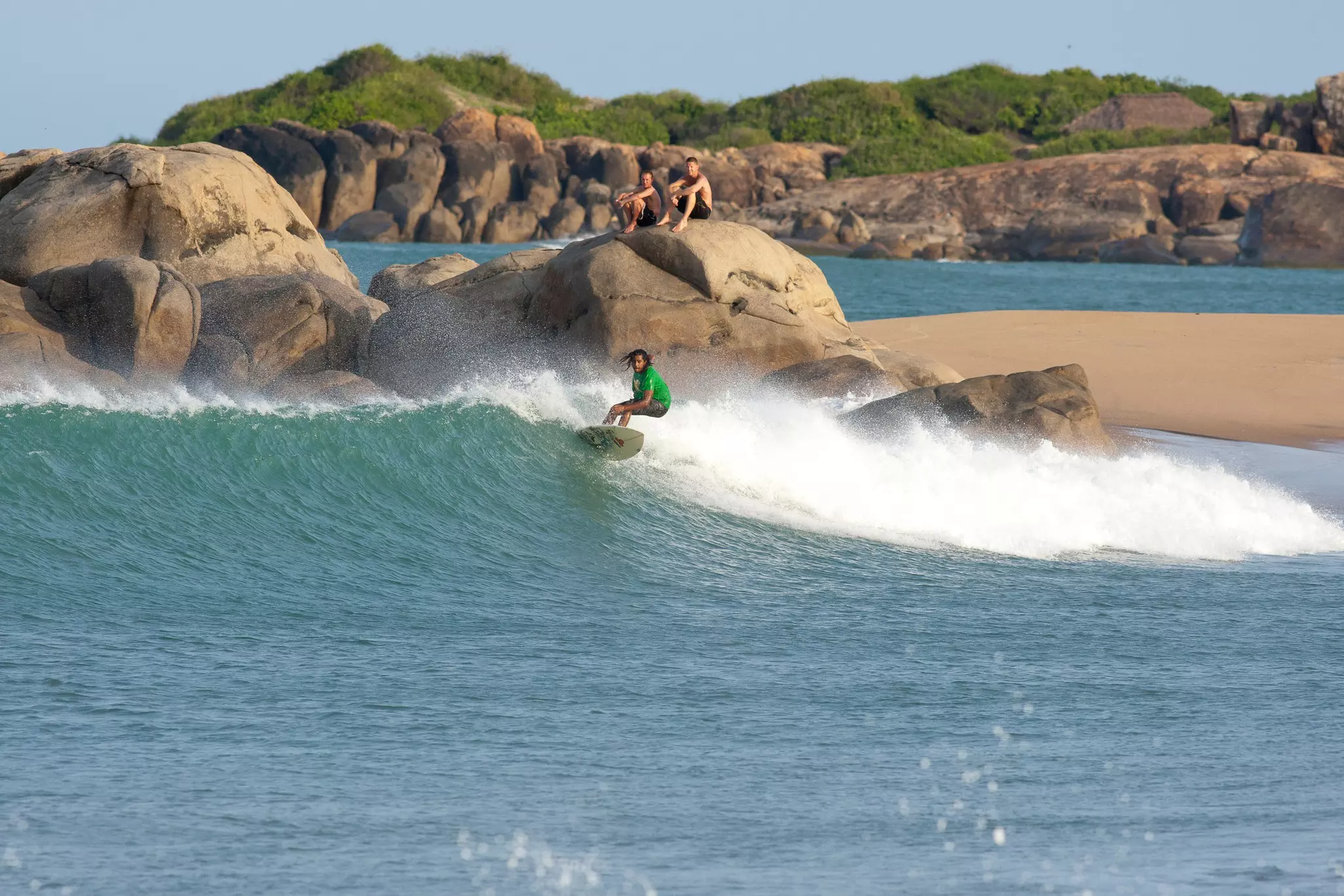 Two people sit on a rock beside the sea watching a surf catch a wave.