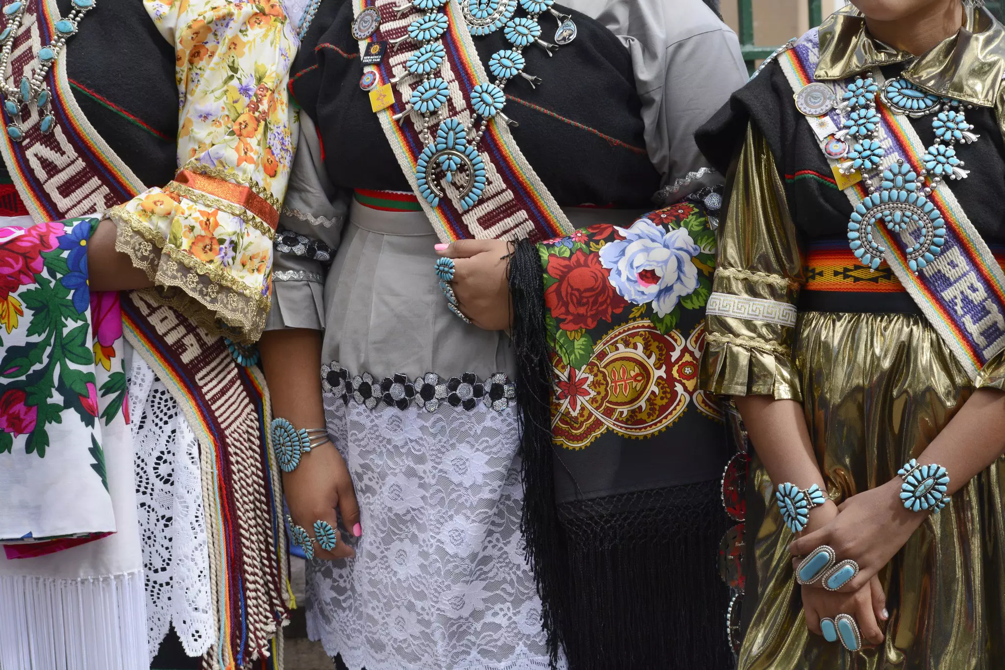 The torsos of three women are seen wearing elaborate outfits, which include beaded sashes, lace skirts and turquoise jewelry.