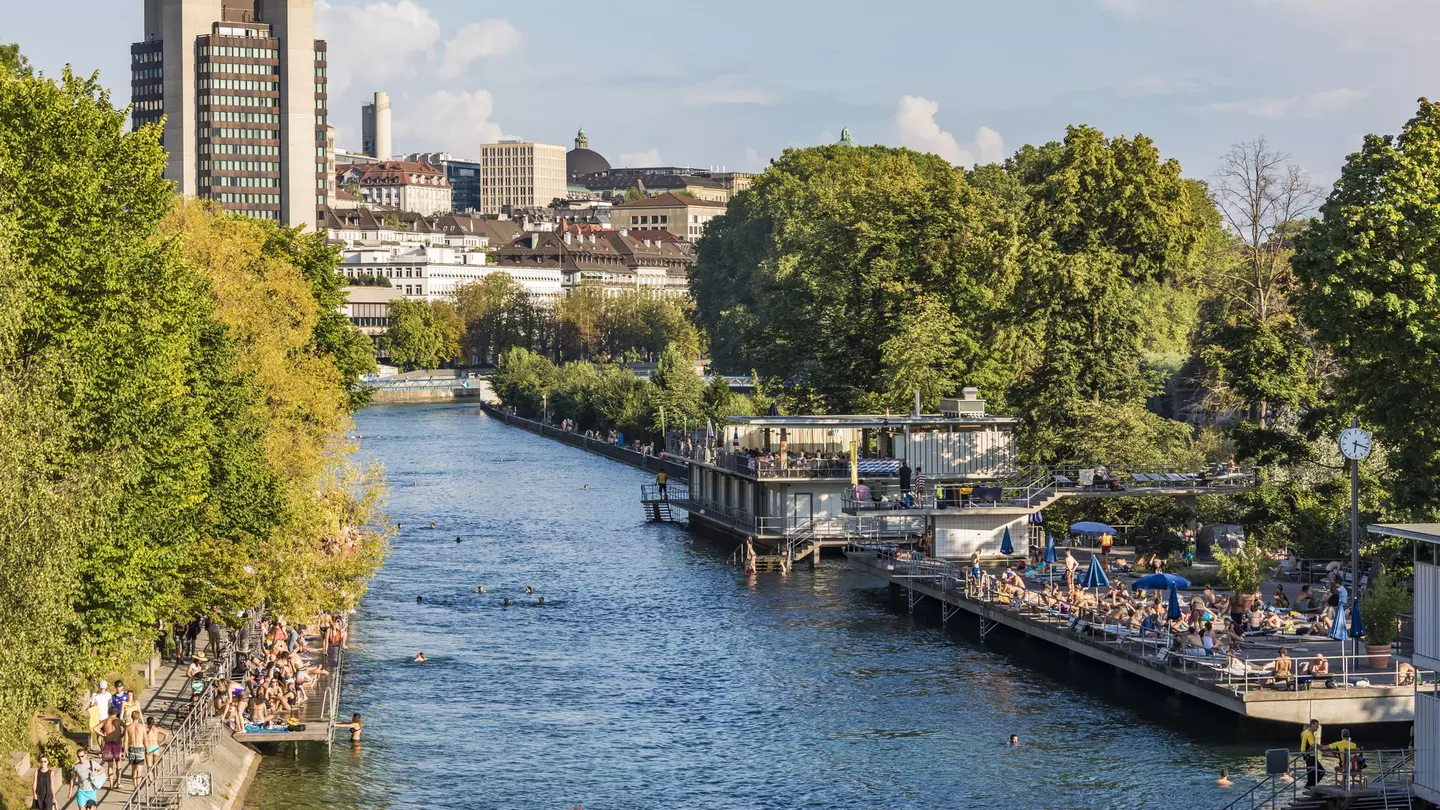 A river running through an urban area. Platforms either side of the river have many people lounging on them, with several swimmers in the river