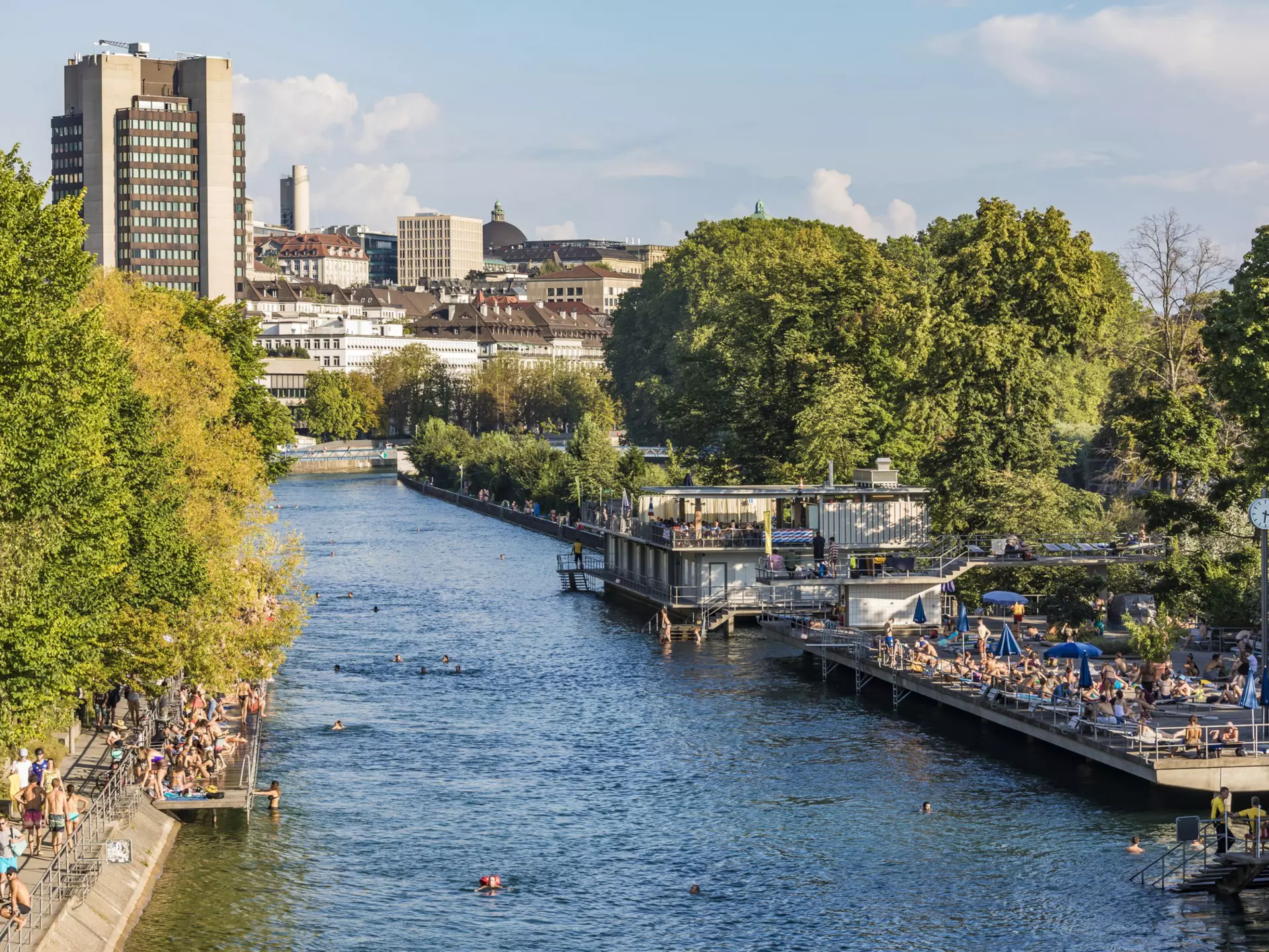 A river running through an urban area. Platforms either side of the river have many people lounging on them, with several swimmers in the river
