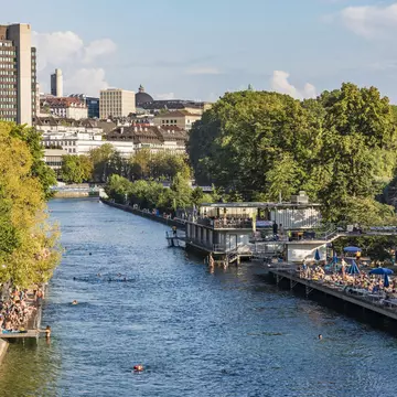 A river running through an urban area. Platforms either side of the river have many people lounging on them, with several swimmers in the river