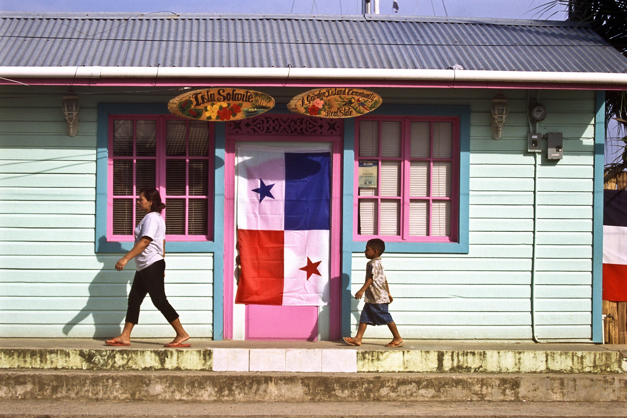 A woman and a small boy walk past a mint-green wooden house with pink shutters and a Panama flag in Bocas Town.