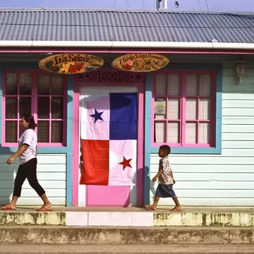 A woman and a small boy walk past a mint-green wooden house with pink shutters and a Panama flag in Bocas Town.