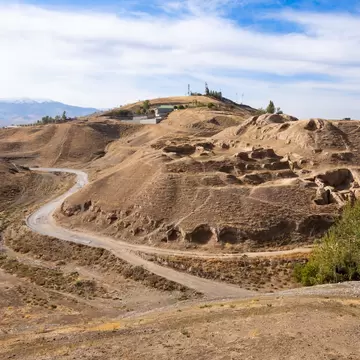 Eroded mud-brick ruins on a dry hillside with a winding dirt road and snowy mountains beyond, Tajikistan