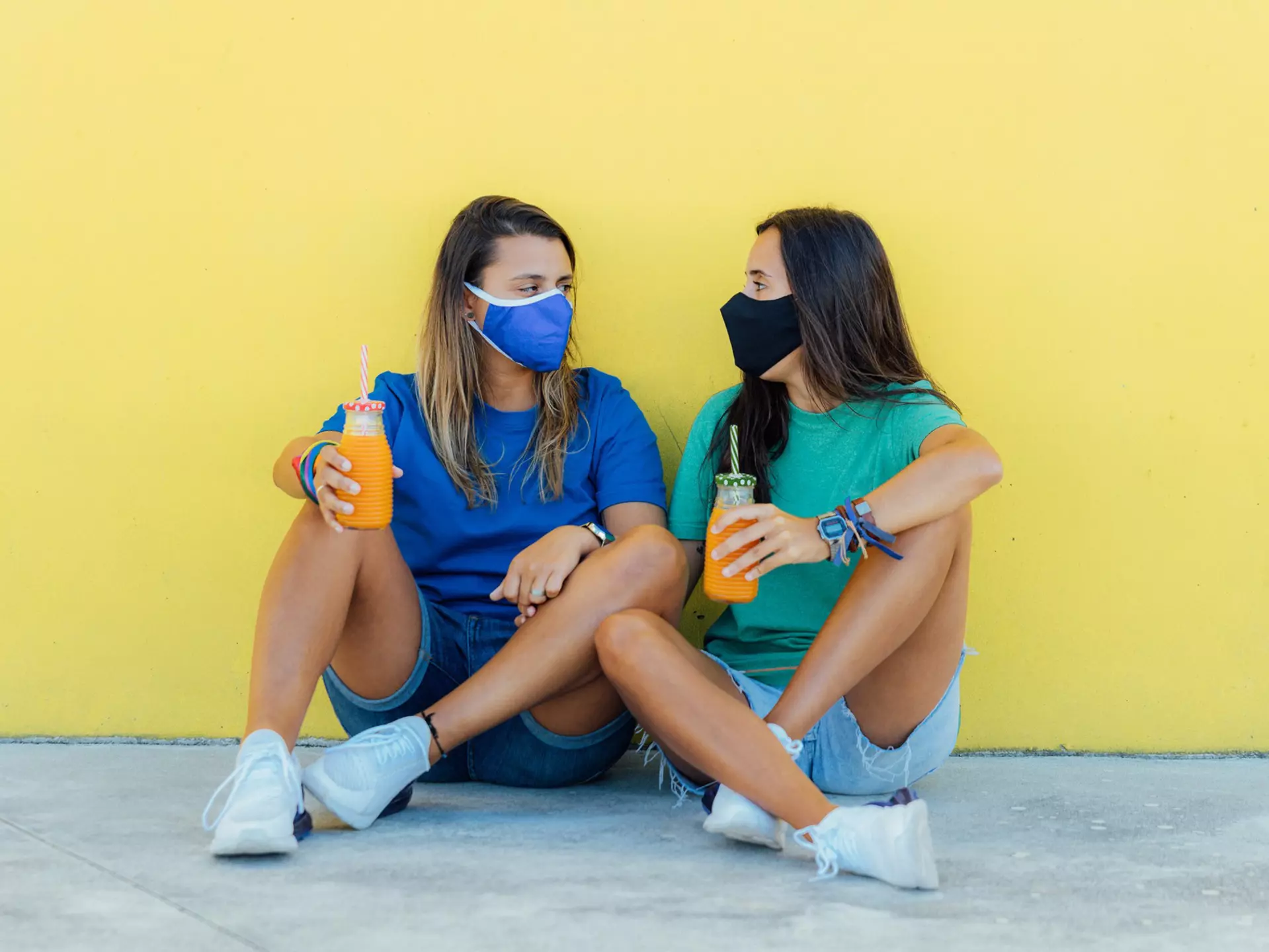 Young lesbian couple sitting wearing medical face masks, holding orange juices with a yellow wall in the background