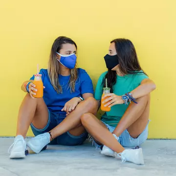 Young lesbian couple sitting wearing medical face masks, holding orange juices with a yellow wall in the background