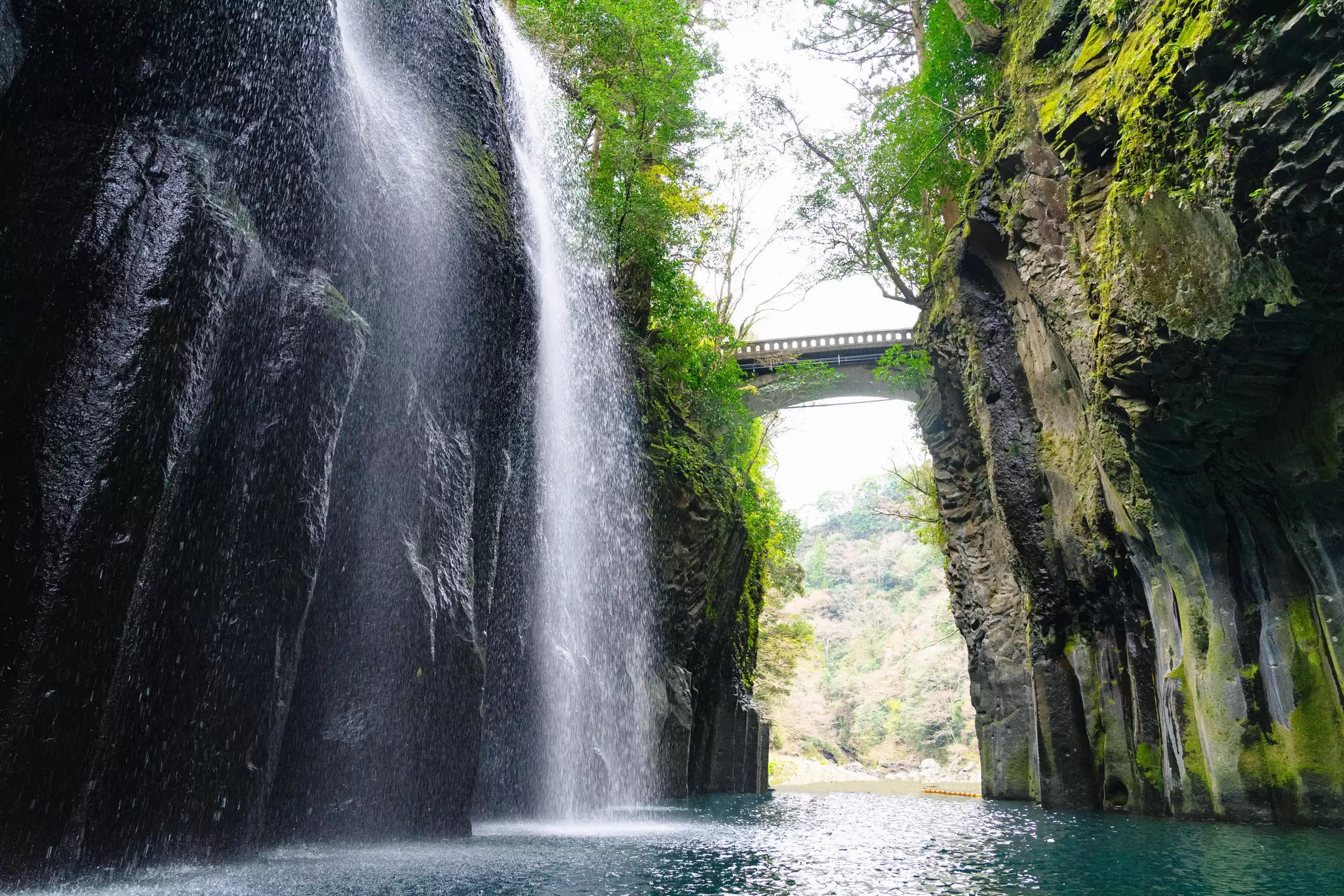 A waterfall down a rocky cliff into a narrow body of water below; an arched bridge spans the distance across the water.