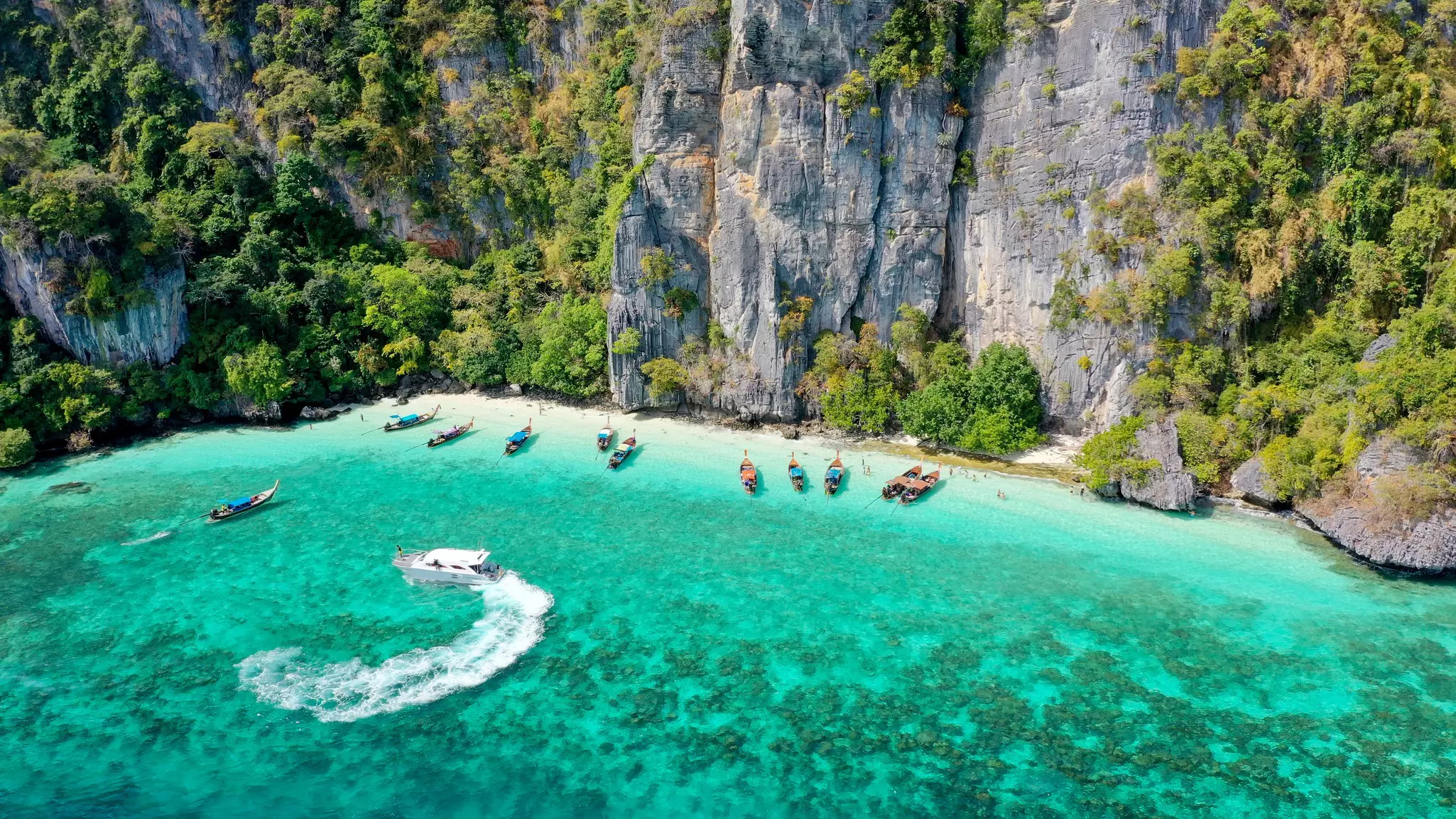 An aerial view of fishing boats and speedboats moored in shallow water next to a cliff.