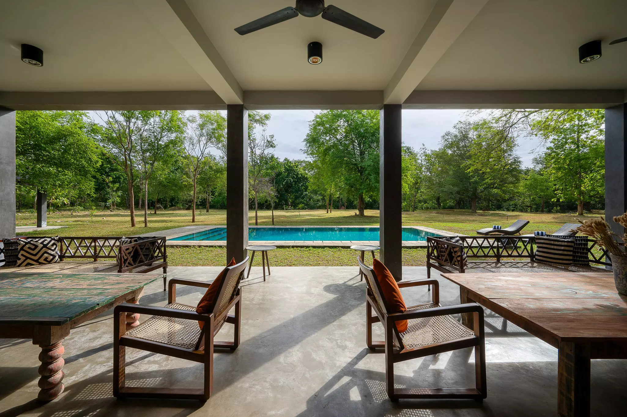 An covered open-fronted terrace with tables and chairs facing a rectangular swimming pool in a large garden with many trees.