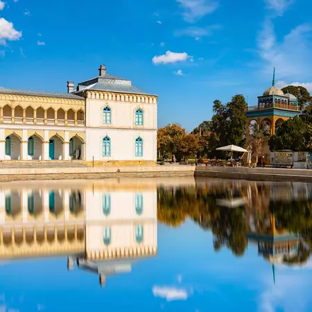 A small palace featuring a loggia with pointed arches is reflected in a pool on a clear day.
