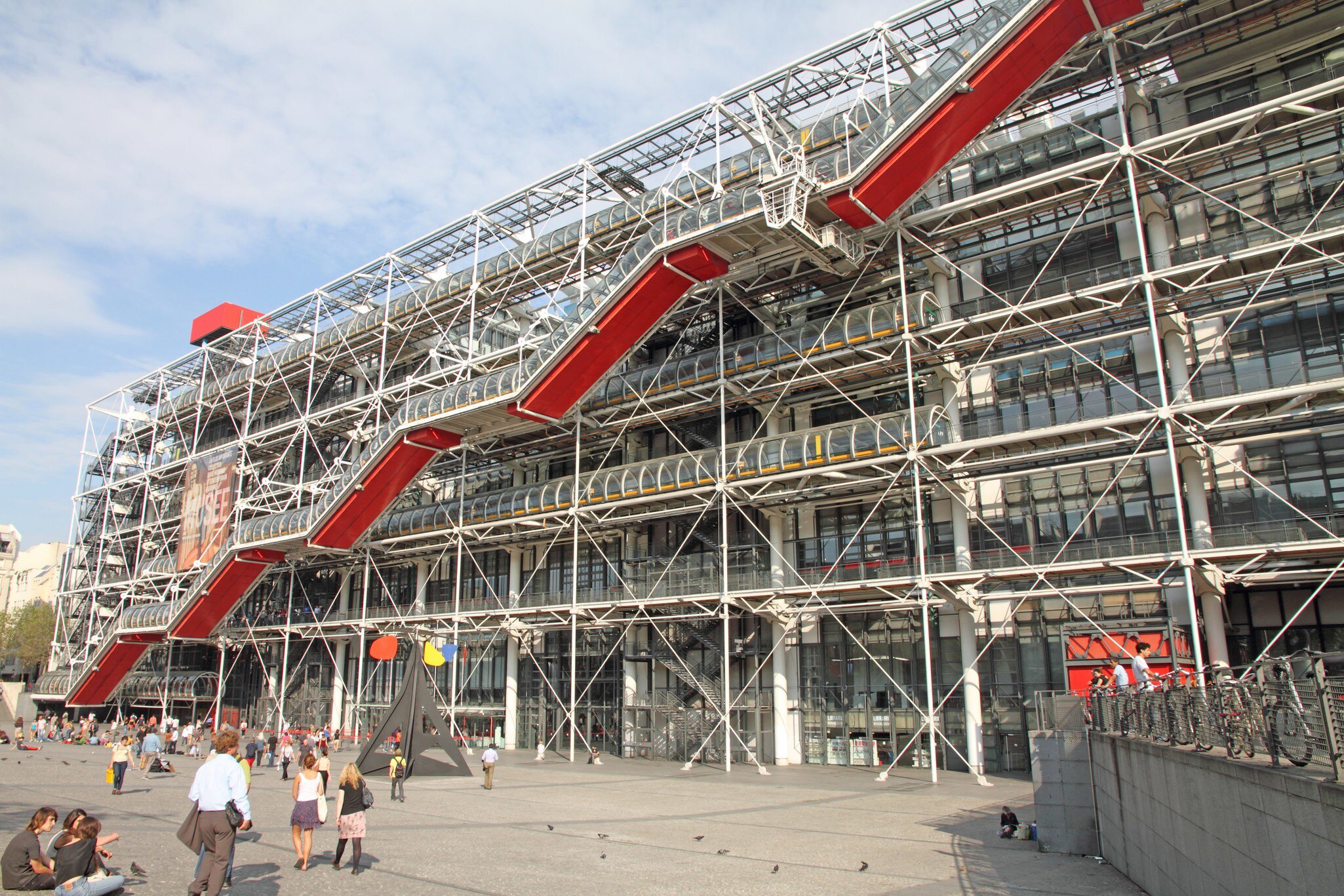 Red escalator tube zigzags up a white steel exoskeleton; a black sculpture with coloured discs sits on the piazza.