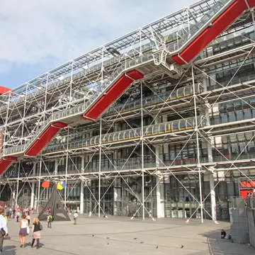 Red escalator tube zigzags up a white steel exoskeleton; a black sculpture with coloured discs sits on the piazza.