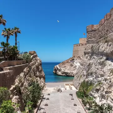 Paved steps lead down between limestone cliffs and crenellated fortress walls to a small beach and deep blue sea.