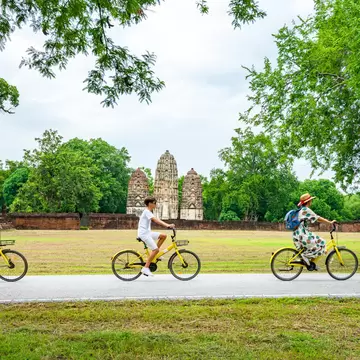 Family cycles on yellow bikes past the three prangs of Wat Si Sawai in Sukhothai Historical Park, Thailand