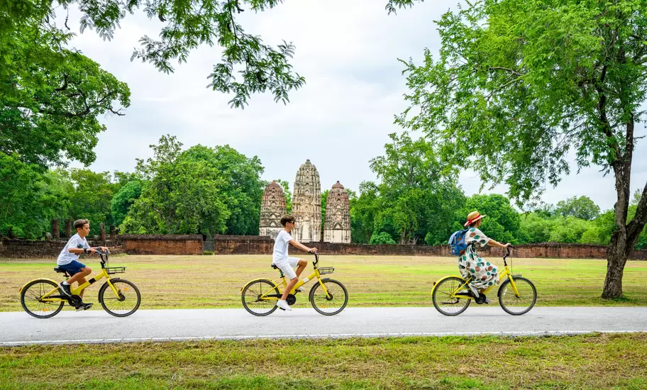 Family cycles on yellow bikes past the three prangs of Wat Si Sawai in Sukhothai Historical Park, Thailand