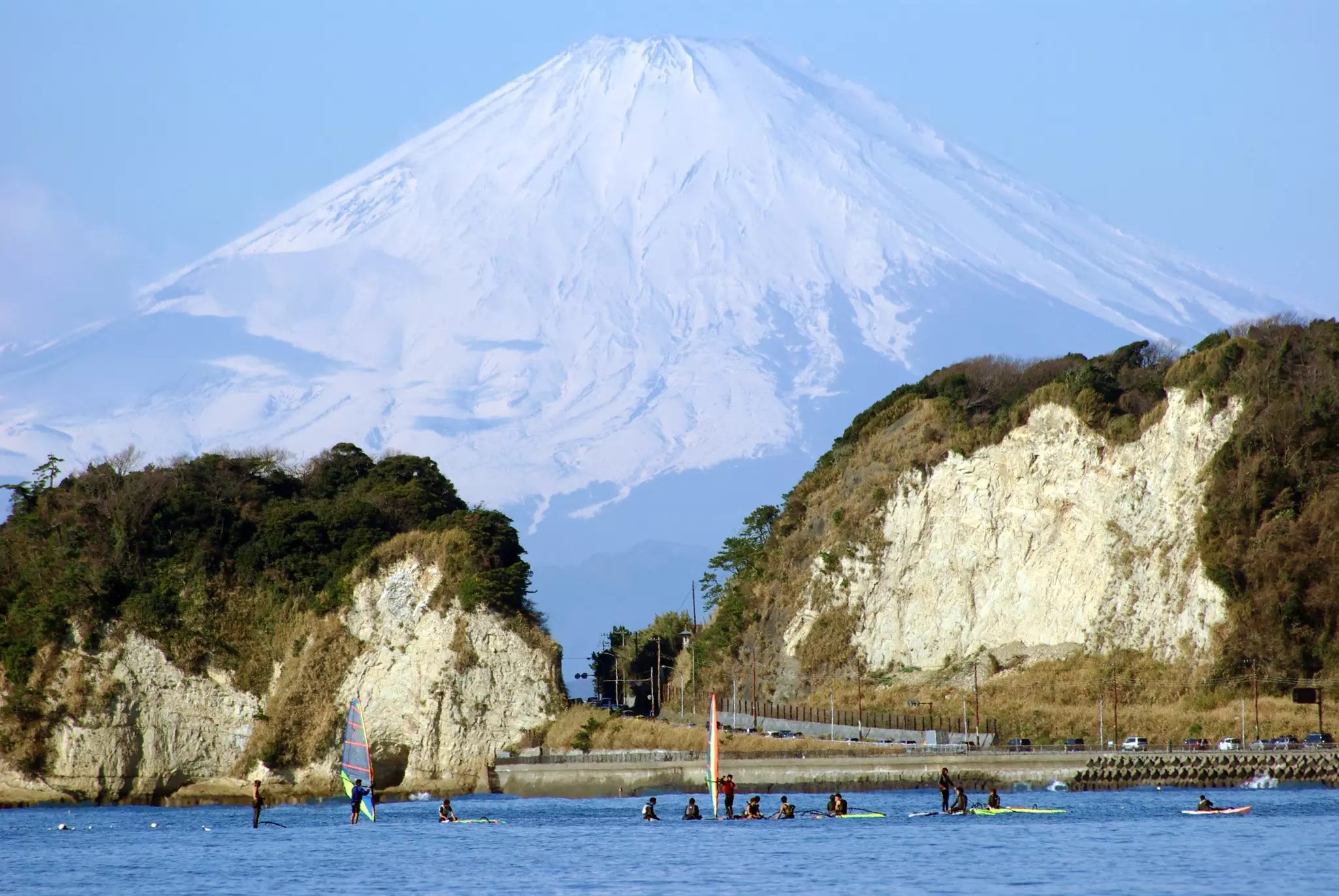 A huge mountain is seen in the distance behind a beach with windsurfers.