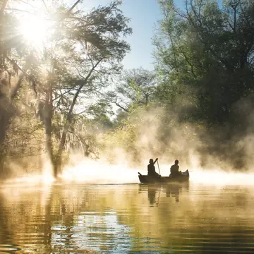 Two people in a canoe are seen in silhouette paddling through a misty river.