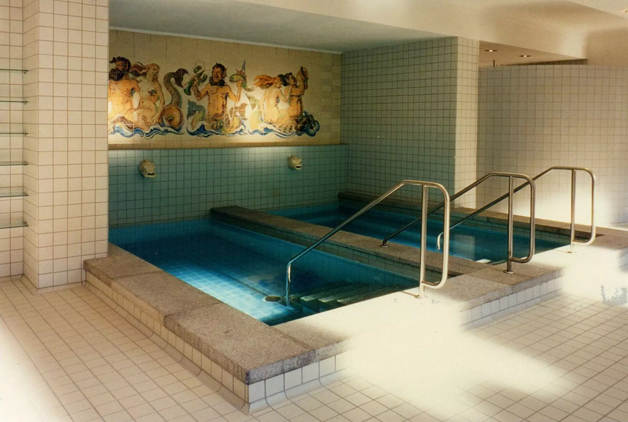 Steps lead down into two small rectangular indoor pools. A mosaic of people at sea is on the wall above.