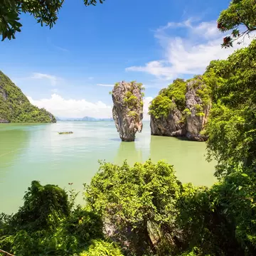 Narrow limestone pinnacle rising from jade-green water beside forested karst islands, a longtail boat nearby