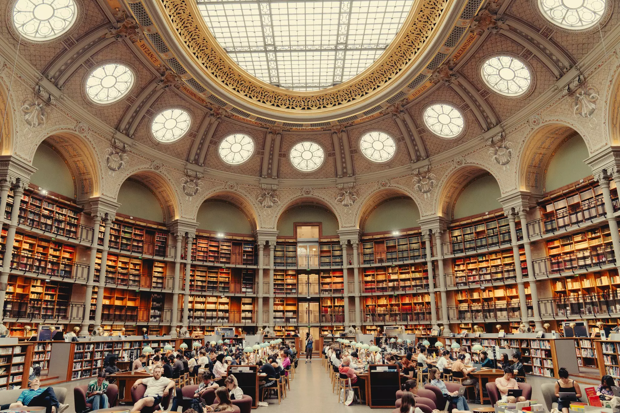 Interior of a vast library with desks for study under an ornate glass ceiling