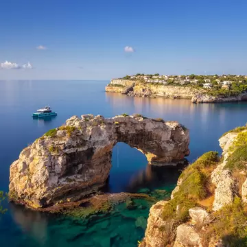 Limestone sea arch in turquoise water off Mallorca's Santanyí coast; a catamaran moored beyond, villas on the headland