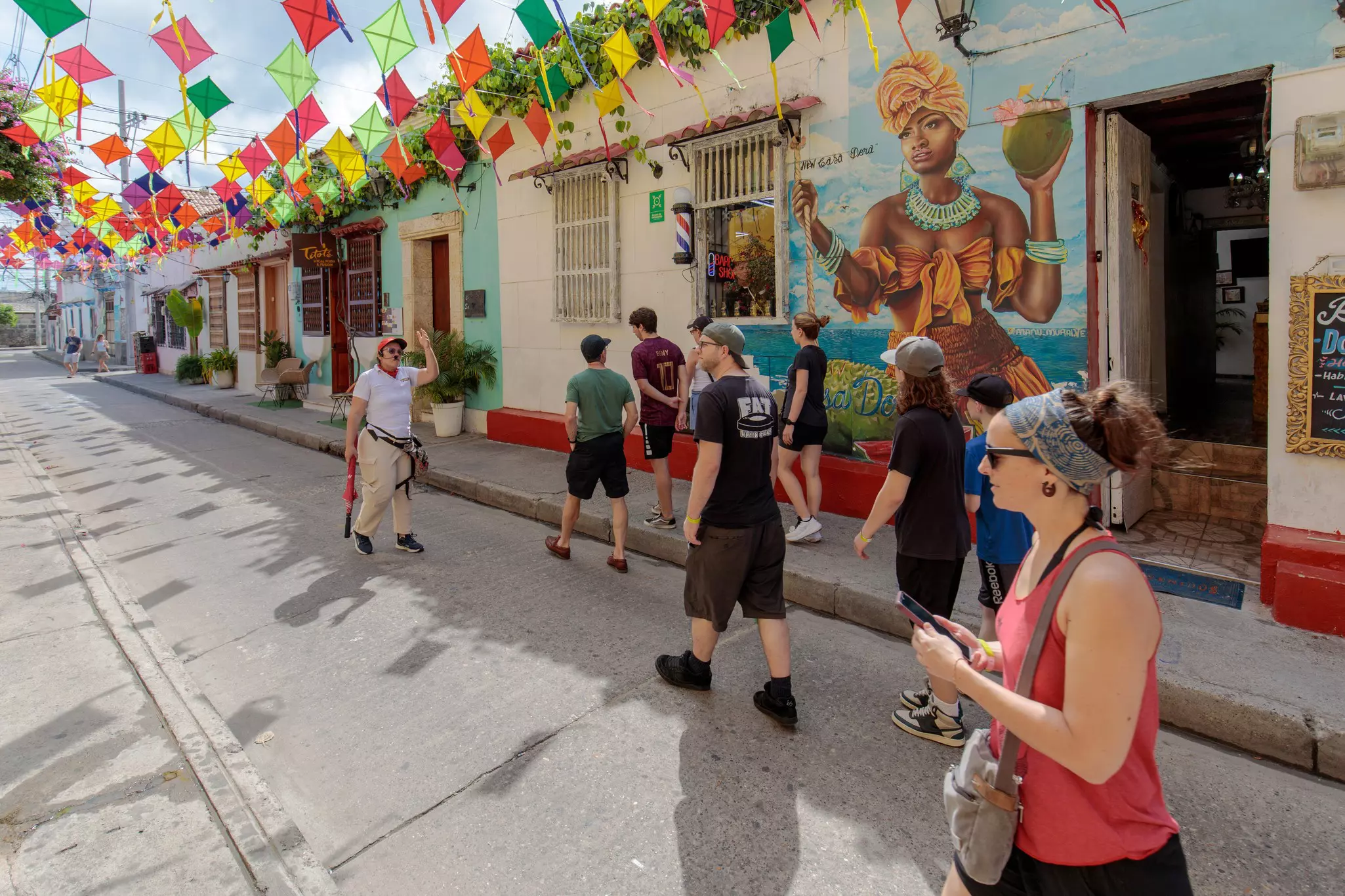 People on a walking tour are pictured on a city street. Paper flags are strewn across the street, and murals decorate the walls of its one-story buildings.