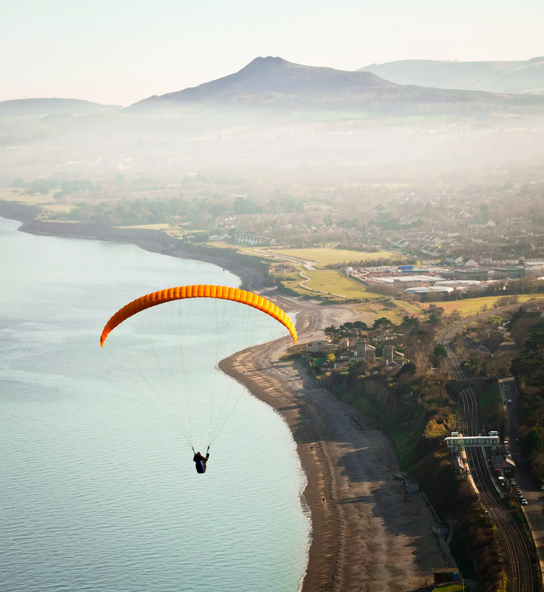  Paragliding descending over coast on his way to beach with sea and mountains in background.