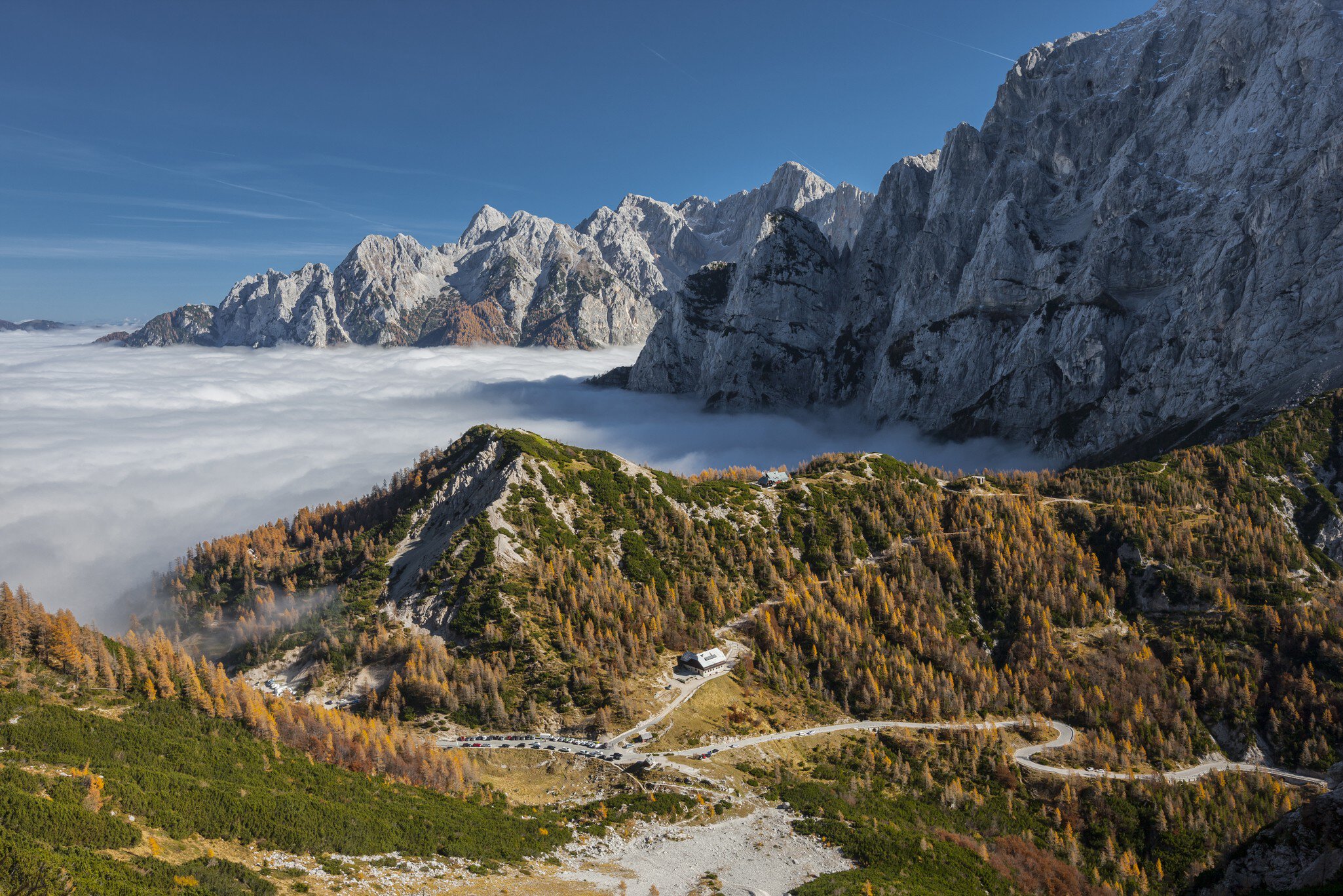Hairpin road through autumn larches above a valley filled with cloud, jagged peaks beyond.