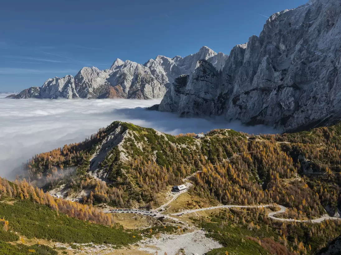 Hairpin road through autumn larches above a valley filled with cloud, jagged peaks beyond.