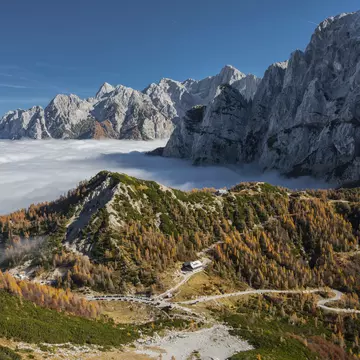 Hairpin road through autumn larches above a valley filled with cloud, jagged peaks beyond.