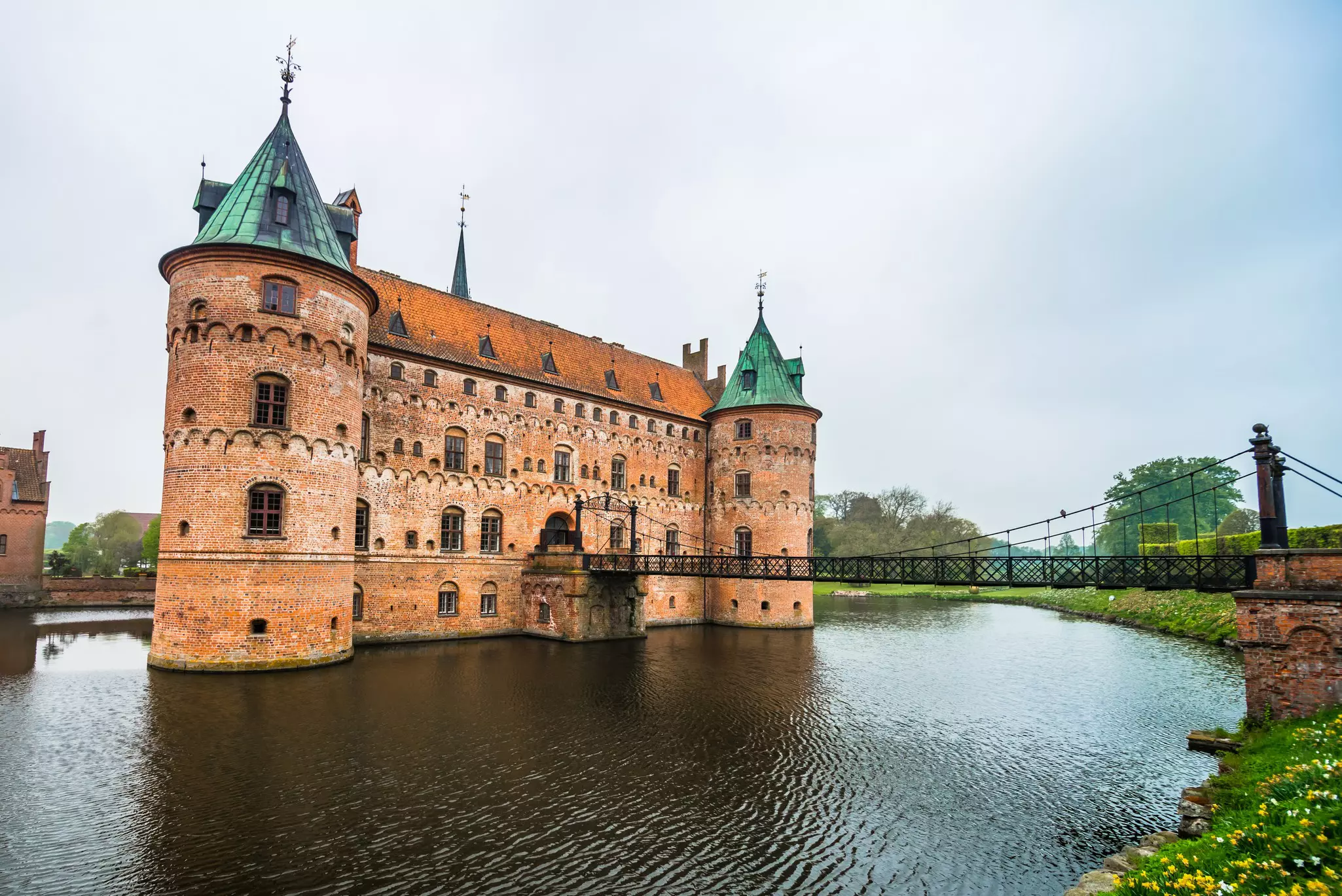 A narrow bridge over a pond leads to an elaborate castle with two hours and a brick facade.