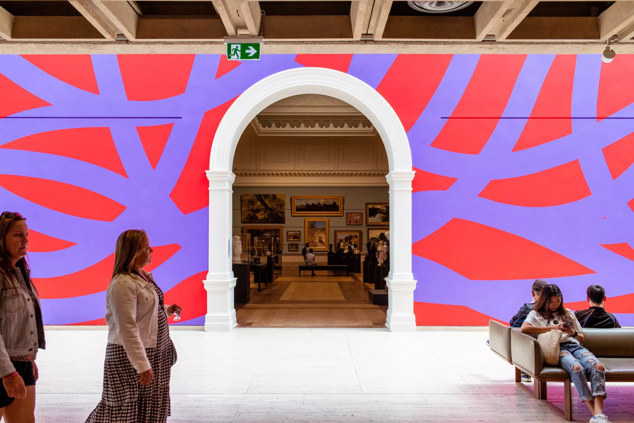 Interior shot of an art gallery with vibrant purple and red walls, a curved entry into a separate gallery and vistors walking through or sitting on benches.