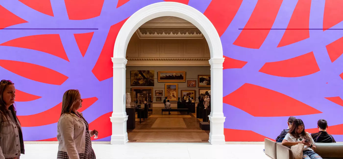 A purple and red patterned wall has a white archway in the center, through which classical paintings are visible.