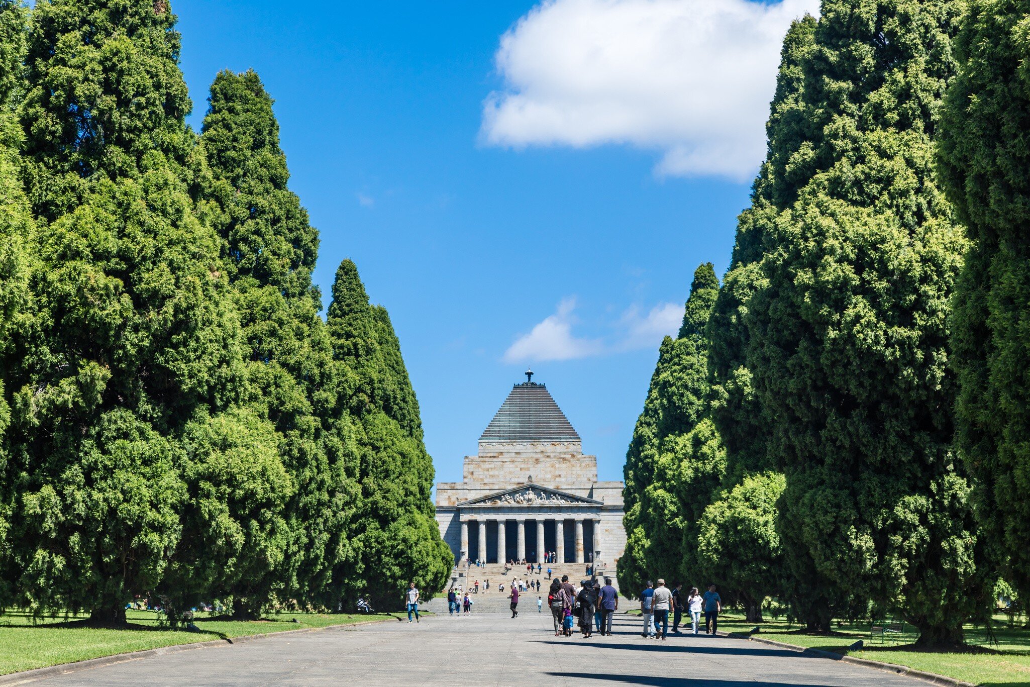 People walking down a tree-lined path towards a large brick memorial.