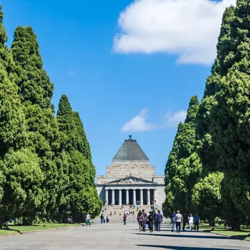 People walking down a tree-lined path towards a large brick memorial.