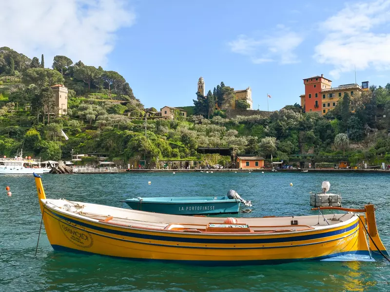 one yellow wooden boat floating on the water with a turquoise boat behind it. The shore is lush green plants with colorful buildings.