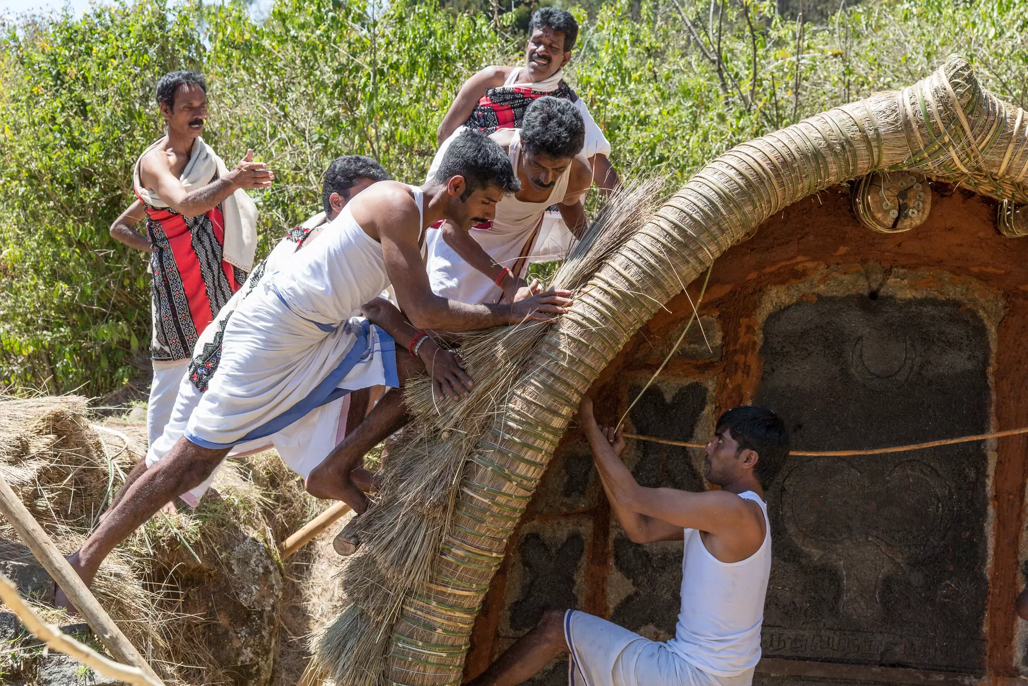 A group of men dressed in white, some with red and black shawls wrapped round them, work together to thatch the roof of a small curved hut.