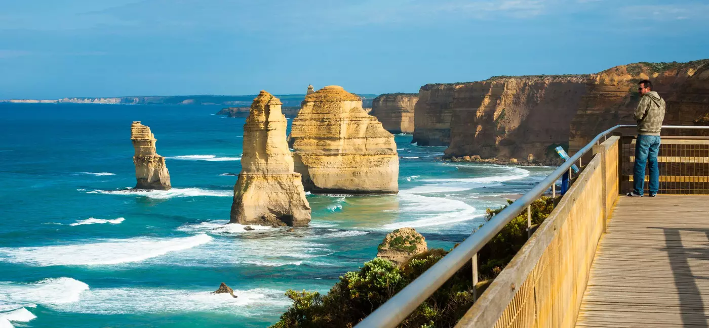 A person on a wooden lookout deck facing rock formations in the ocean off the shore from tall cliffs.