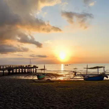 Outrigger fishing boats pulled up on dark sand beside a wooden pier, with the sun low over the sea under golden clouds.