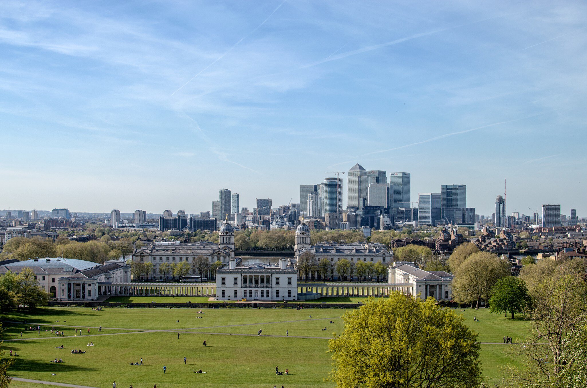 People scattered on a wide lawn above colonnaded white buildings and a distant cluster of towers.