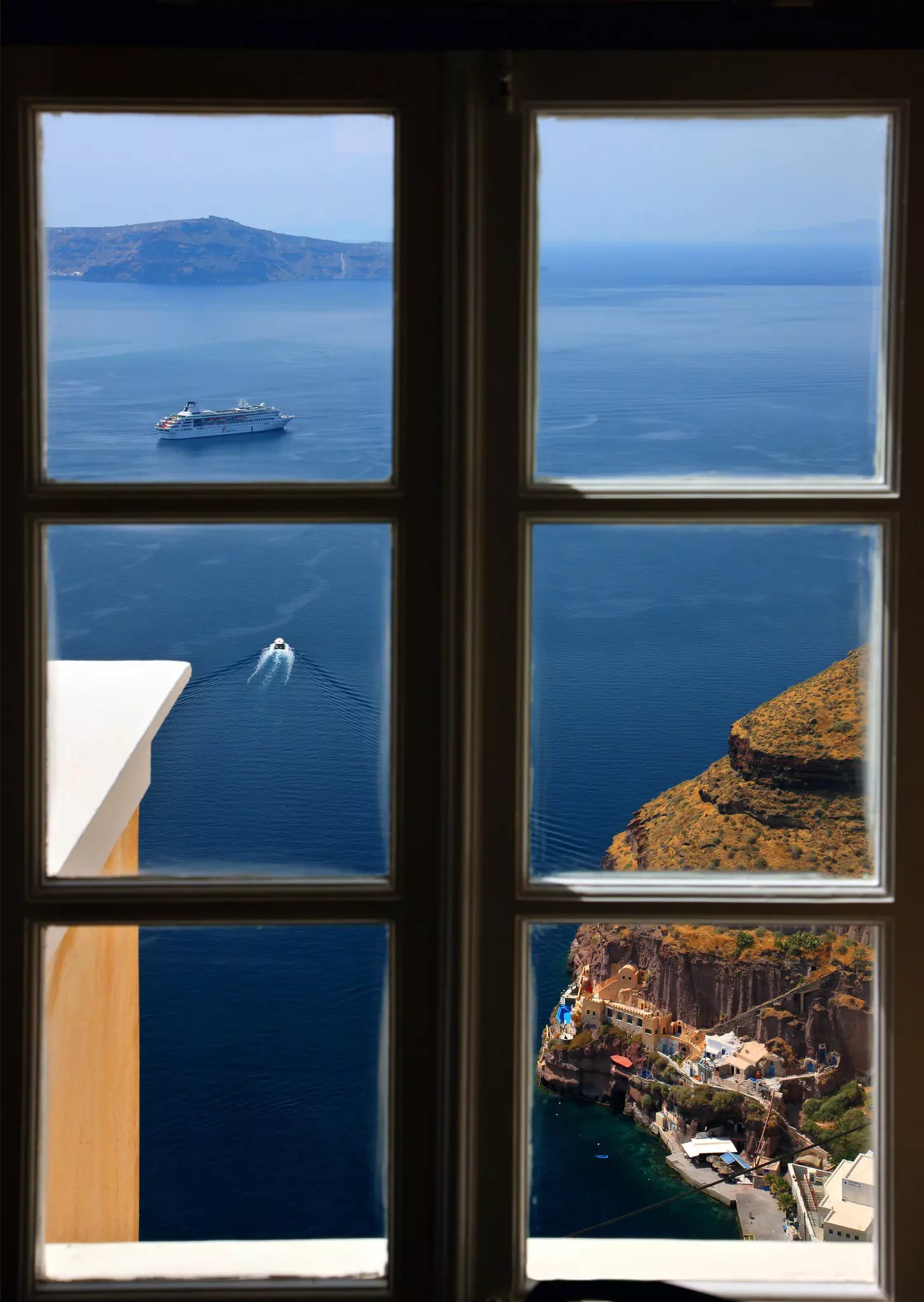 A view of a caldera through a window in a Greek village with grassy cliffs and colorful buildings in the pic.