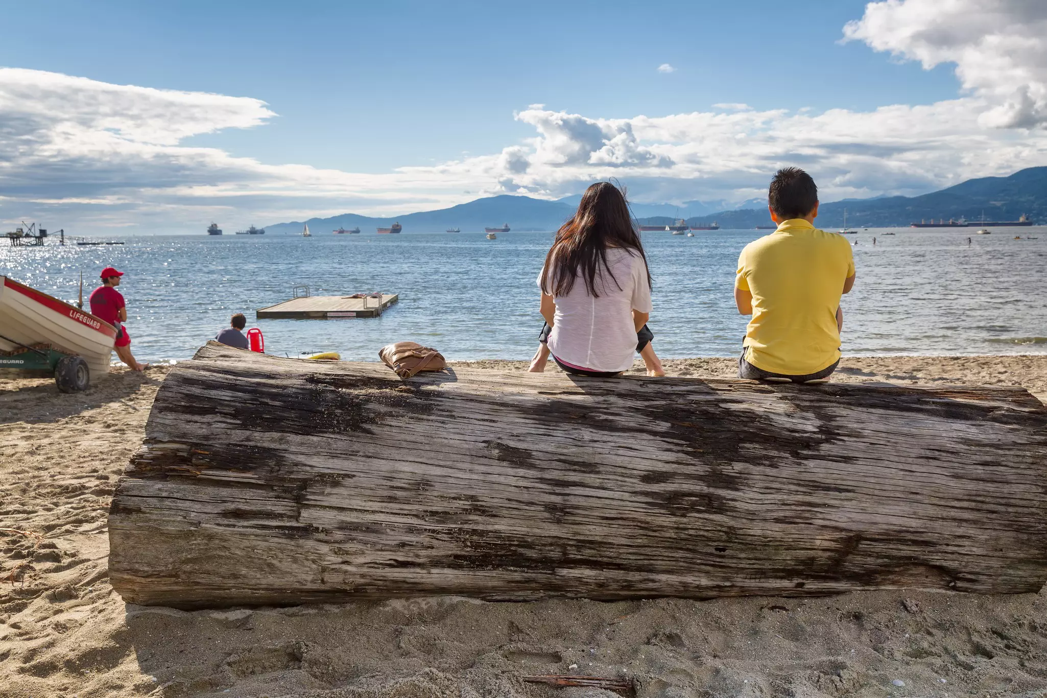 Two people sit on a large driftwood log on a sandy beach looking at the water in Vancouver, Canada.