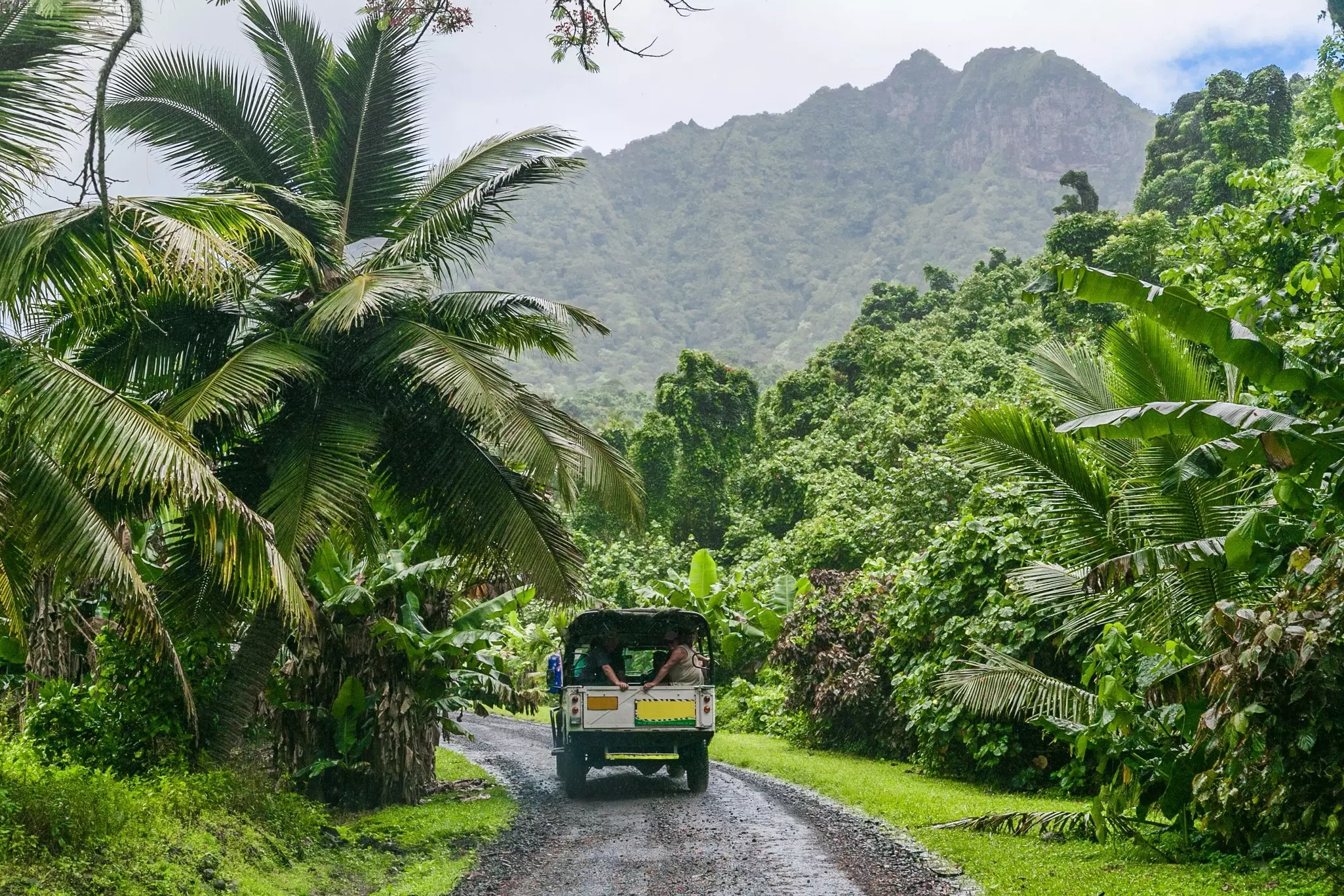 An open Jeep with passengers drives down a dirt road in a lush tropical landscape. Verdant mountains rise in the distance.